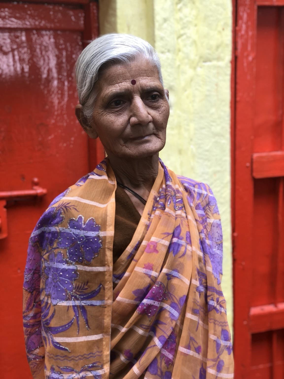 An elderly woman in a patterned sari stands before vivid red doors in Varanasi, 2019