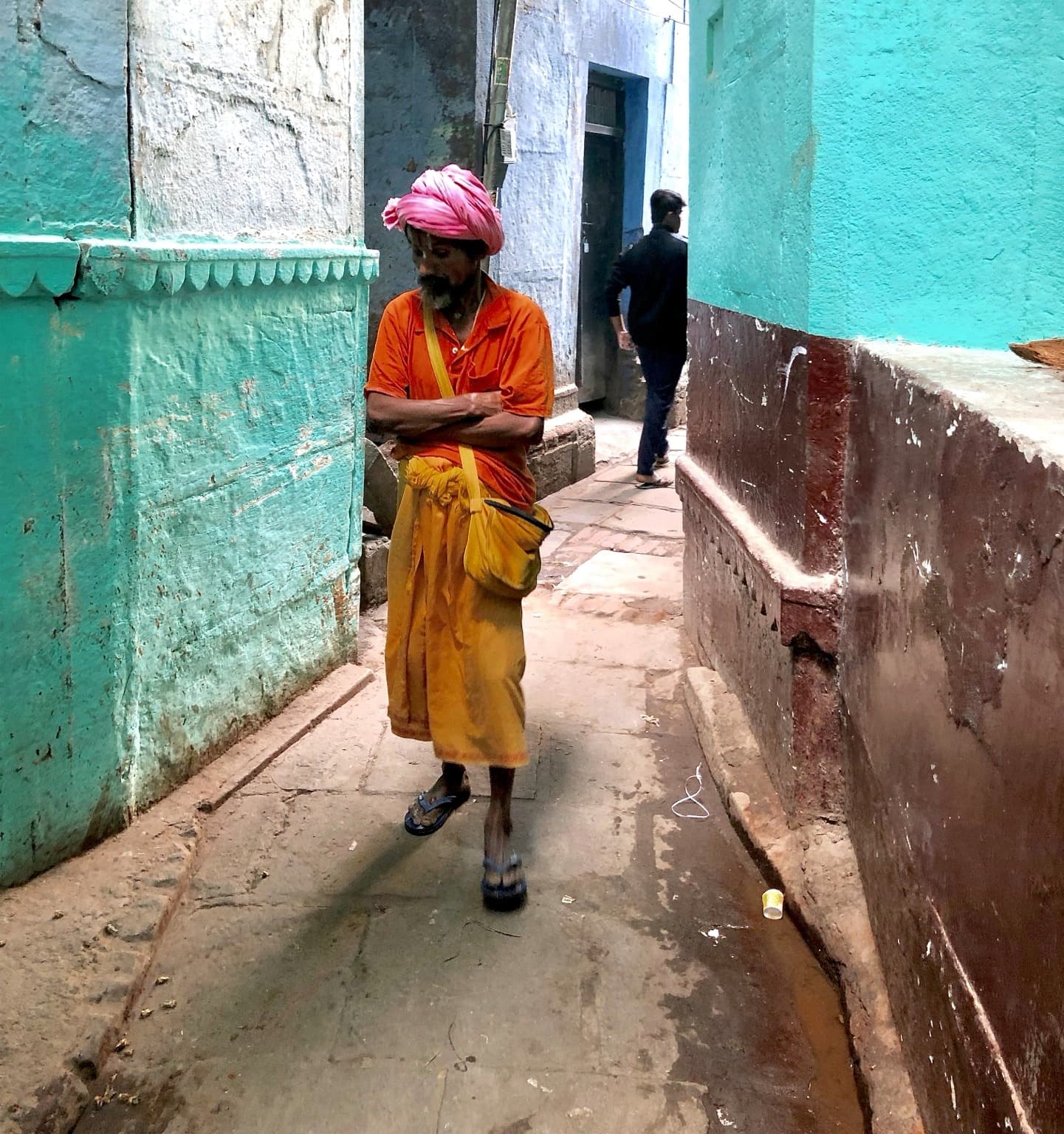 A man in a pink turban, orange shirt and yellow cloth walks through a turquoise-painted lane in Varanasi, 2019