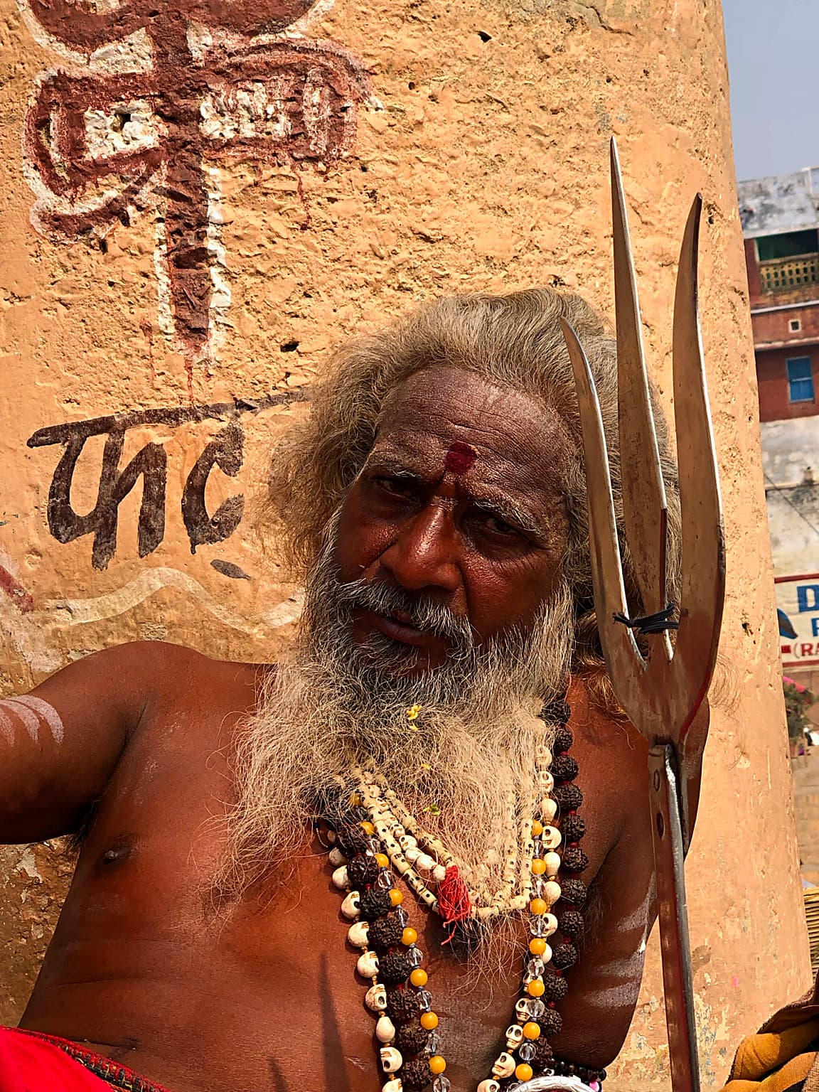 Closer portrait of the gray-haired sadhu with beads and trident beside a textured wall.
