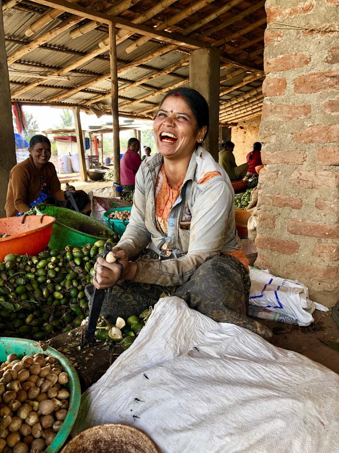 Woman seated among vegetables laughing while holding a knife and produce, with another smiling worker beside her.