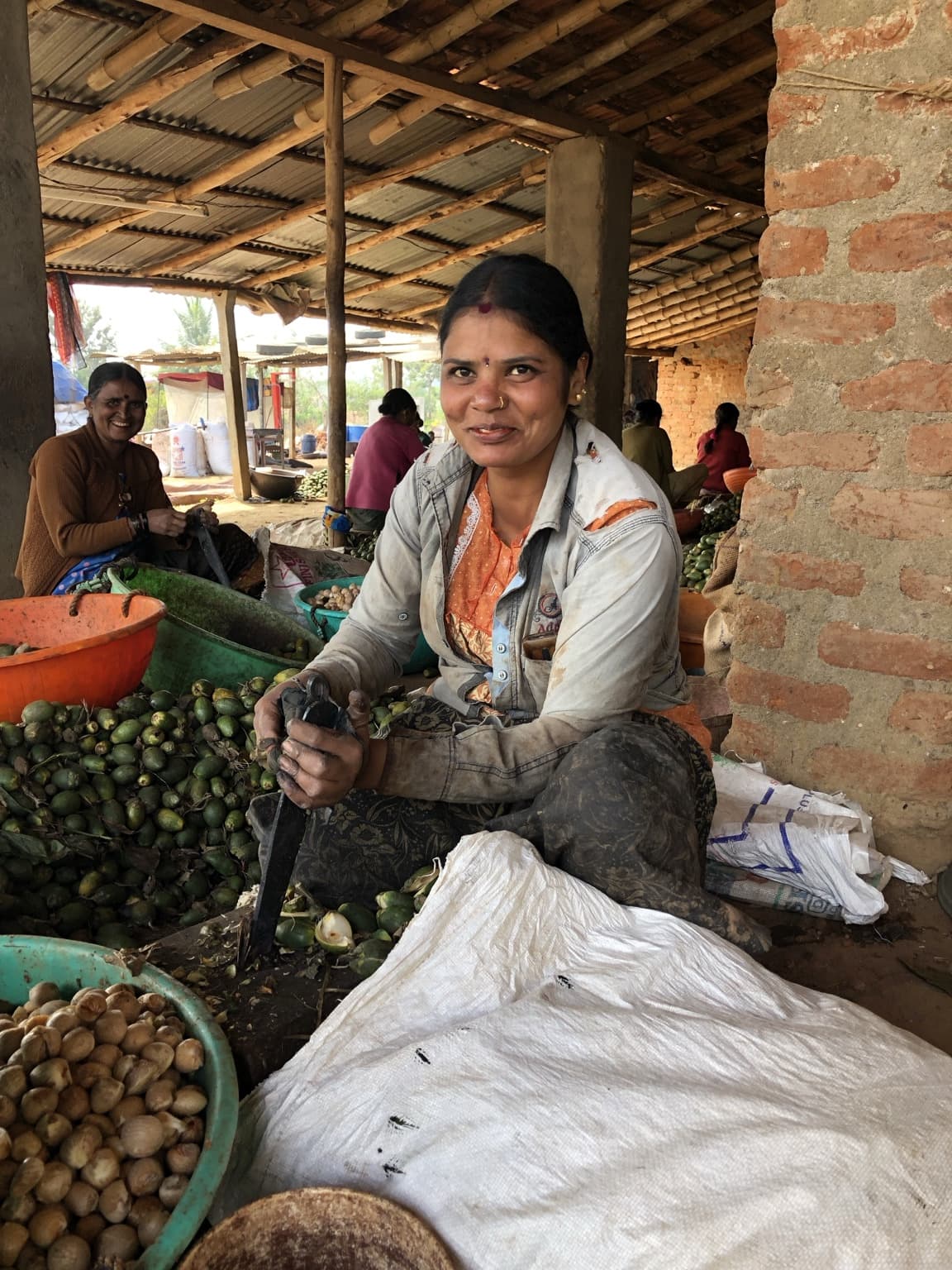Woman looking toward the camera while seated among produce and containers under a market roof.