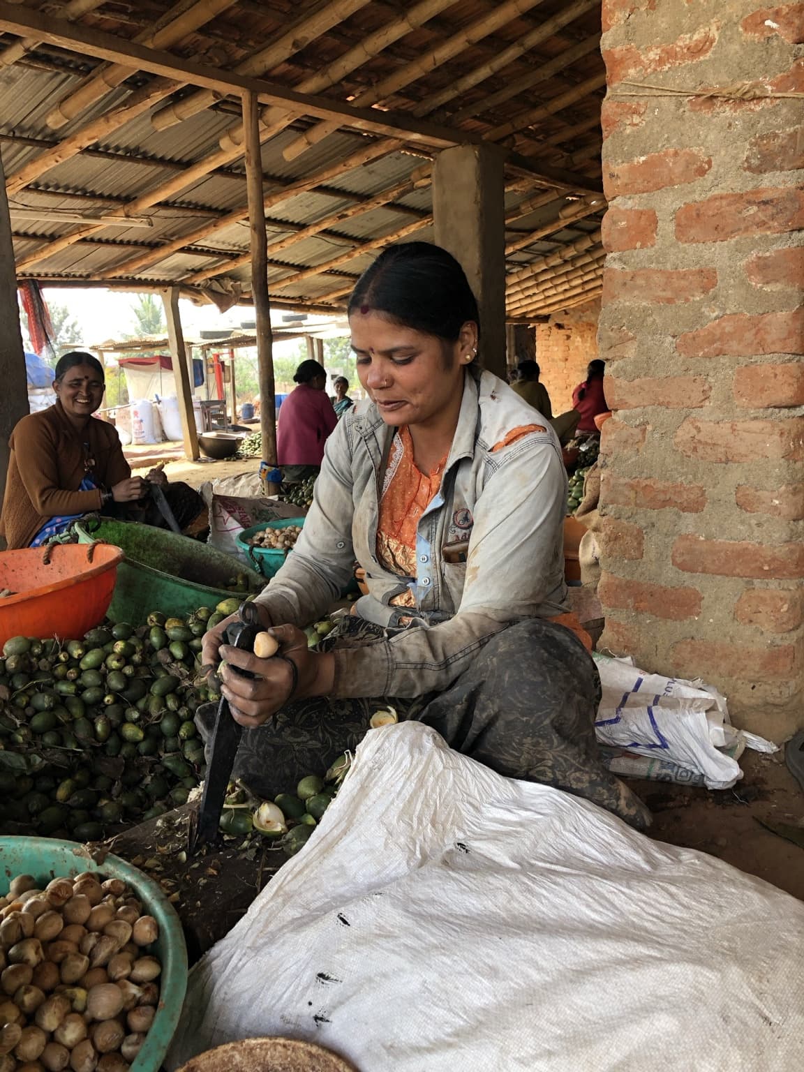 Woman looking down at the produce she is preparing, seated among vegetables and containers beside a brick pillar.