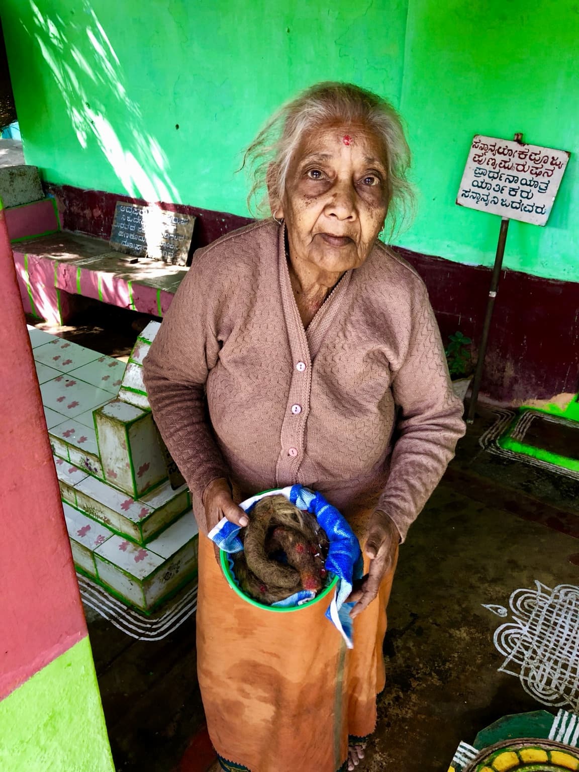 Woman holding a green bowl containing the long hair she had preserved, inside a brightly painted doorway.