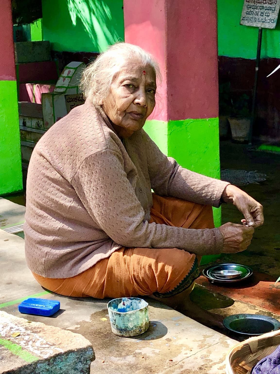Elderly woman seated beside bowls in a brightly painted green and pink doorway.