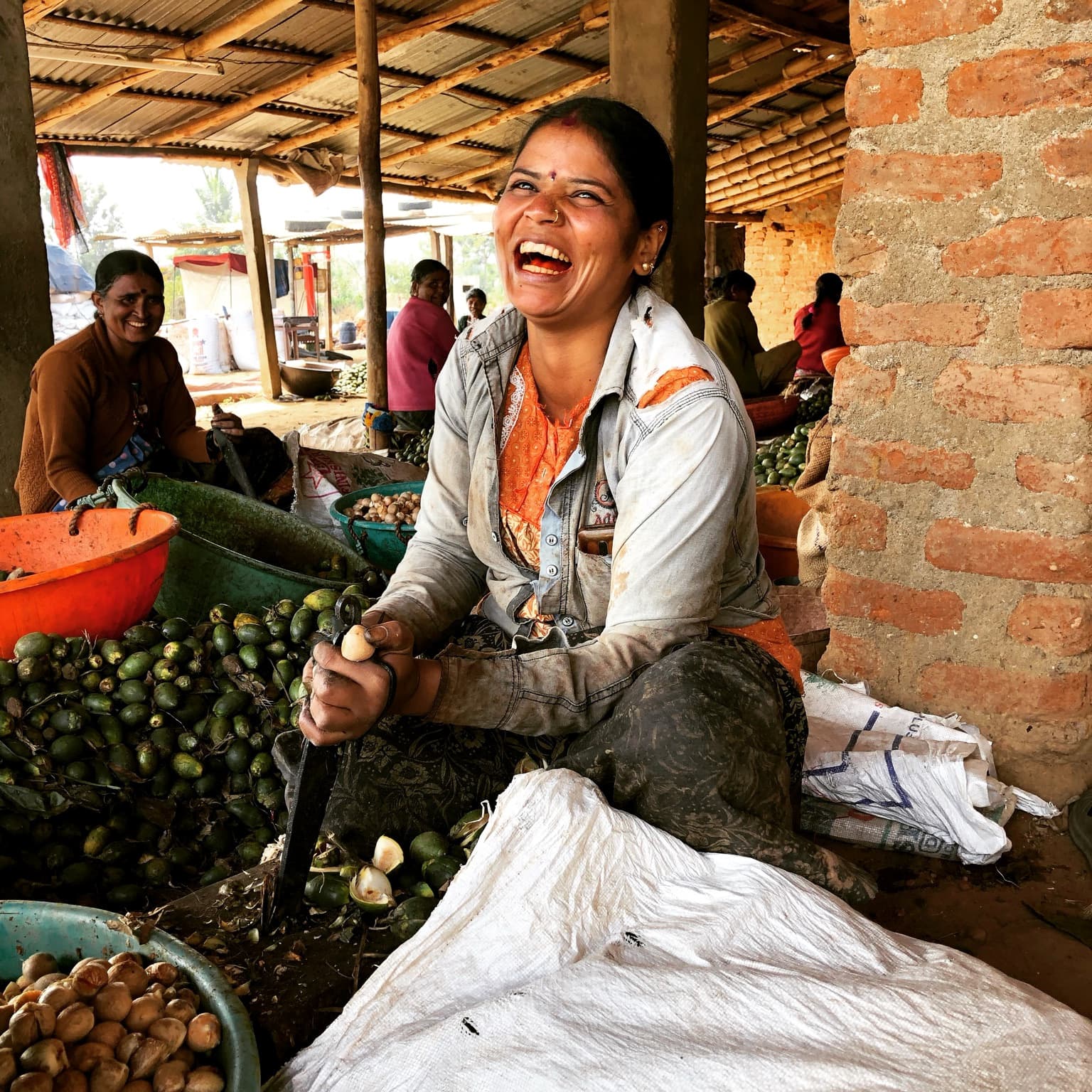 Closer view of the woman laughing at work, with produce, sacks and a brick pillar still visible around her.
