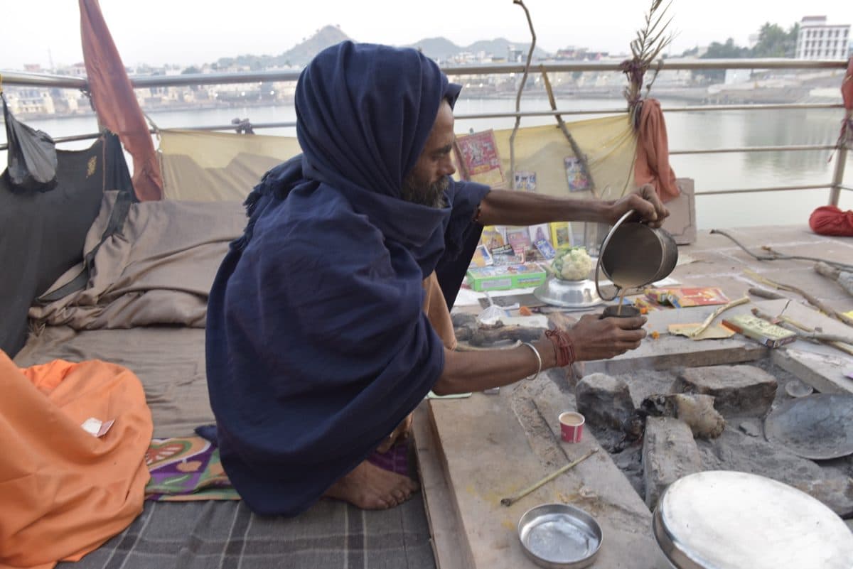 Man in a blue shawl seated beside cooking vessels at Pushkar Lake.